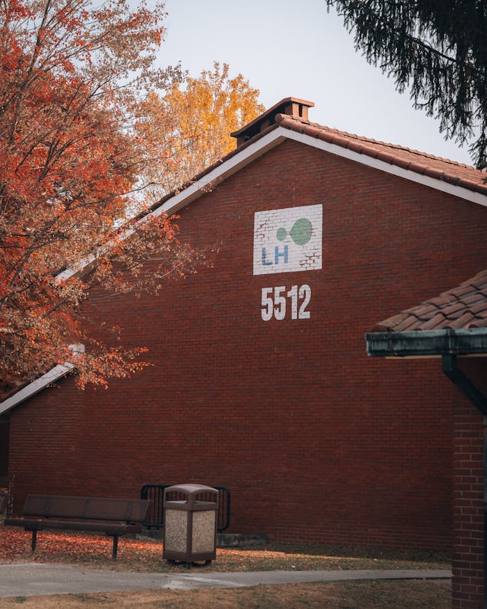 Red brick building in fall, with trees and a bench, featuring signage.